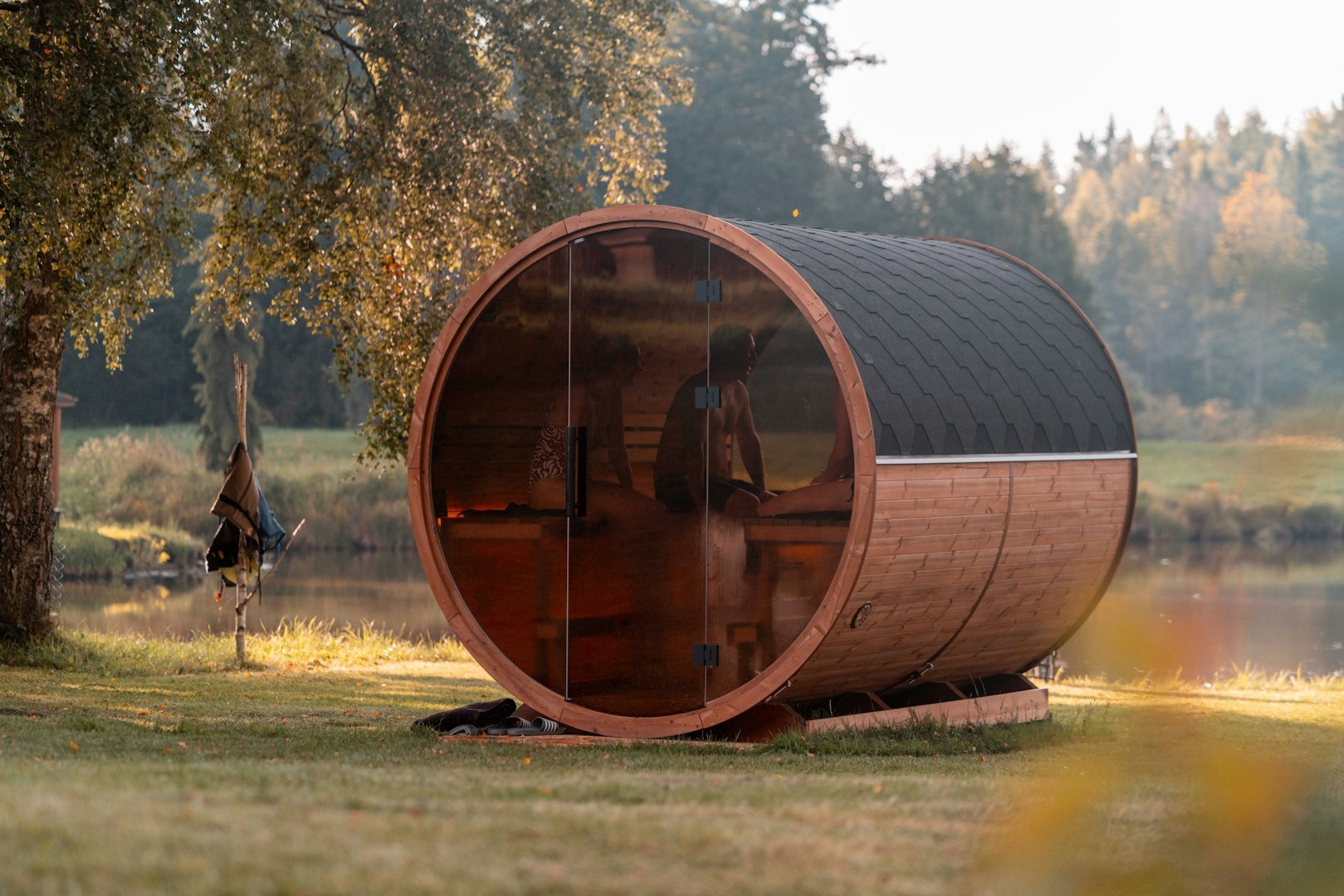 Drewniana sauna beczkowa z przeszklonym frontem, ustawiona nad wodą wśród drzew